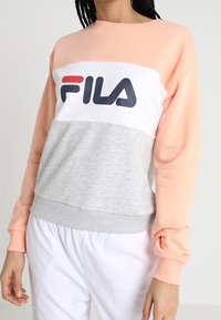 Persoon draagt een color-block FILA sweatshirt in perzik, wit en grijs, gecombineerd met losse witte broek, staand tegen een effen achtergrond.