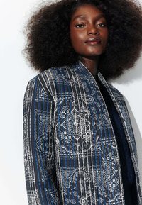 Veste matelassée noire et bleue, ornée de motifs blancs complexes. Texture douce avec un col mandarin et des détails de couture subtils.