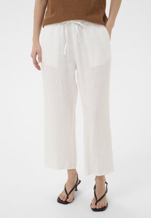 Žena oblečená v voľných bielych ľanových nohaviciach so šnúrkou v páse a čiernymi sandálmi na opätku, stojaca s rukami vo vreckách.