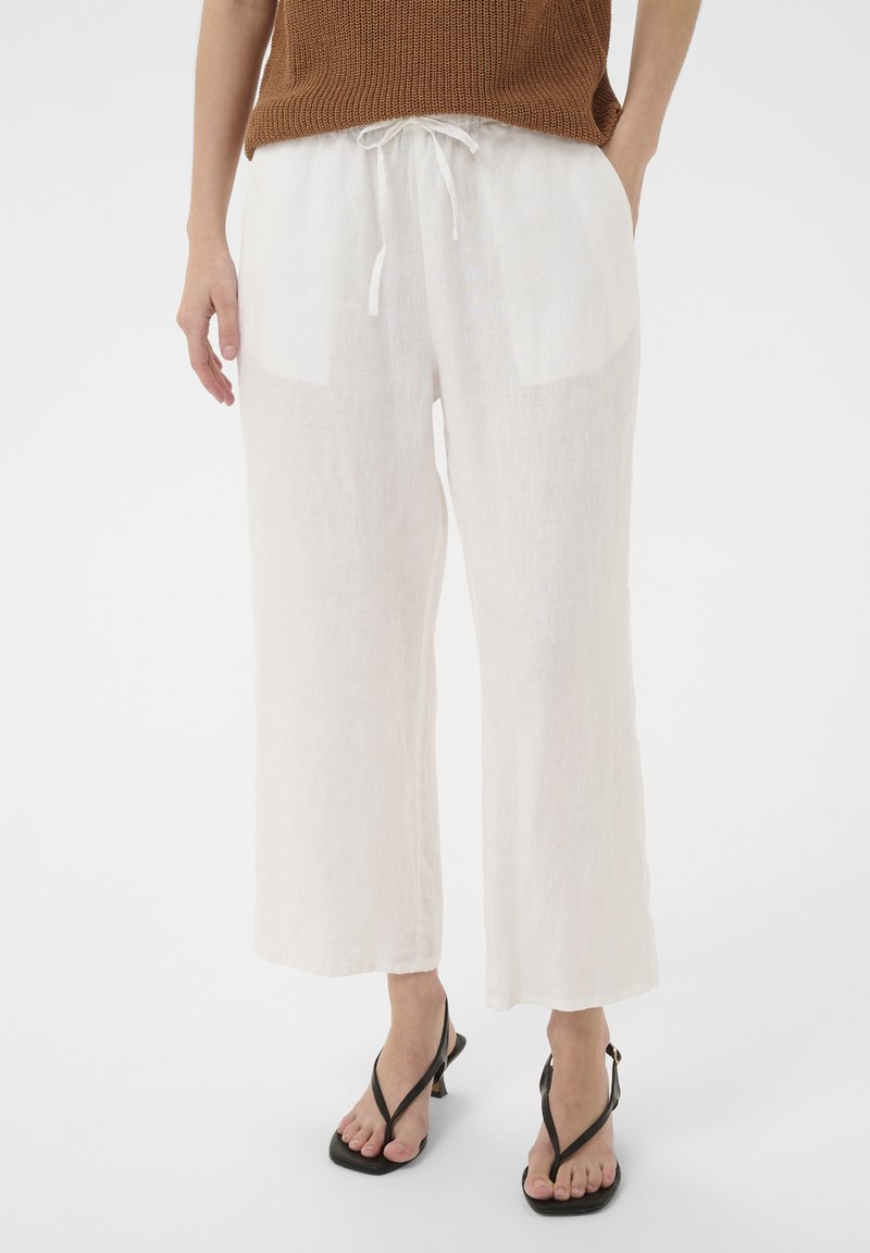 Femme portant un pantalon en lin blanc ample avec taille à cordon de serrage et des sandales à talons noires, debout les mains dans les poches.