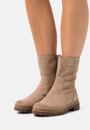 Personne portant des bottes en suède beige mi-mollet avec fermeture éclair latérale et semelle en caoutchouc épaisse, debout devant un fond clair uni.
