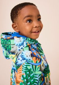 Colorful hooded rain jacket featuring a vibrant jungle pattern with dinosaurs and tropical leaves against a blue background.