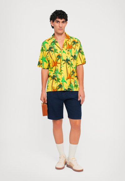 Kurzärmeliges gelbes Hemd mit Palmenmotiv, marineblaue Shorts, cremefarbene Socken und beigefarbene Schuhe. Kleine Tasche am Hemd, die eine braune Tasche hält.
