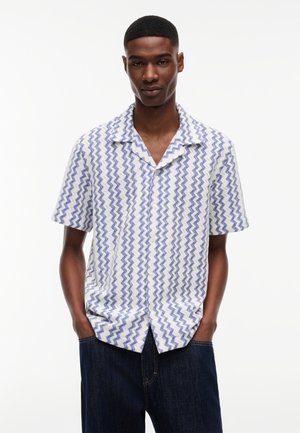 Jonge man met kort haar die een wit overhemd met korte mouwen draagt met blauwe zigzagpatronen en een donkere spijkerbroek, handen in de zakken.