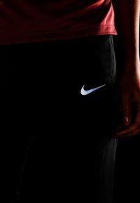 Czarne spodnie sportowe z białym logo Nike, połączone z czerwonym topem. Spodnie mają gładką teksturę i subtelny perforowany wzór w okolicy kostki.