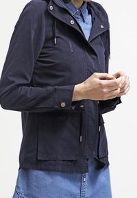 Granatowa kurtka wykonana z lekkiego materiału, wyposażona w kaptur, przednie kieszenie oraz sznurki ściągające. Gładka tekstura z metalicznymi akcentami.