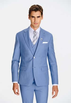 Mężczyzna w jasnoniebieskim garniturze z dopasowaną kamizelką, białą koszulą, szarą krawatem w paski i kwiatową broszką na klapie, stojący na jednolitym tle.