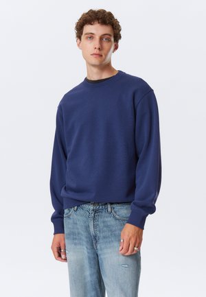 Navy blue sweatshirt with a classic crew neck, long sleeves, and a relaxed fit, paired with faded blue jeans. Smooth texture, minimal design.