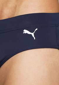 Jūras zila peldkostīma apakša, izgatavota no gludas auduma, ar baltu Puma logotipu; zema jostasvieta un minimālas šuves komfortam.