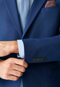 Tengerészkék szövetes blézer texturált kivitelben, kék ingmandzsetta és világoskék mintás nyakkendő. Az ujjon négy fekete gomb található.