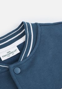 Granatowy sweter z ribbowanym kołnierzem z białymi paskami, z przodu zapinany na napy, z metką materiałową wewnątrz.