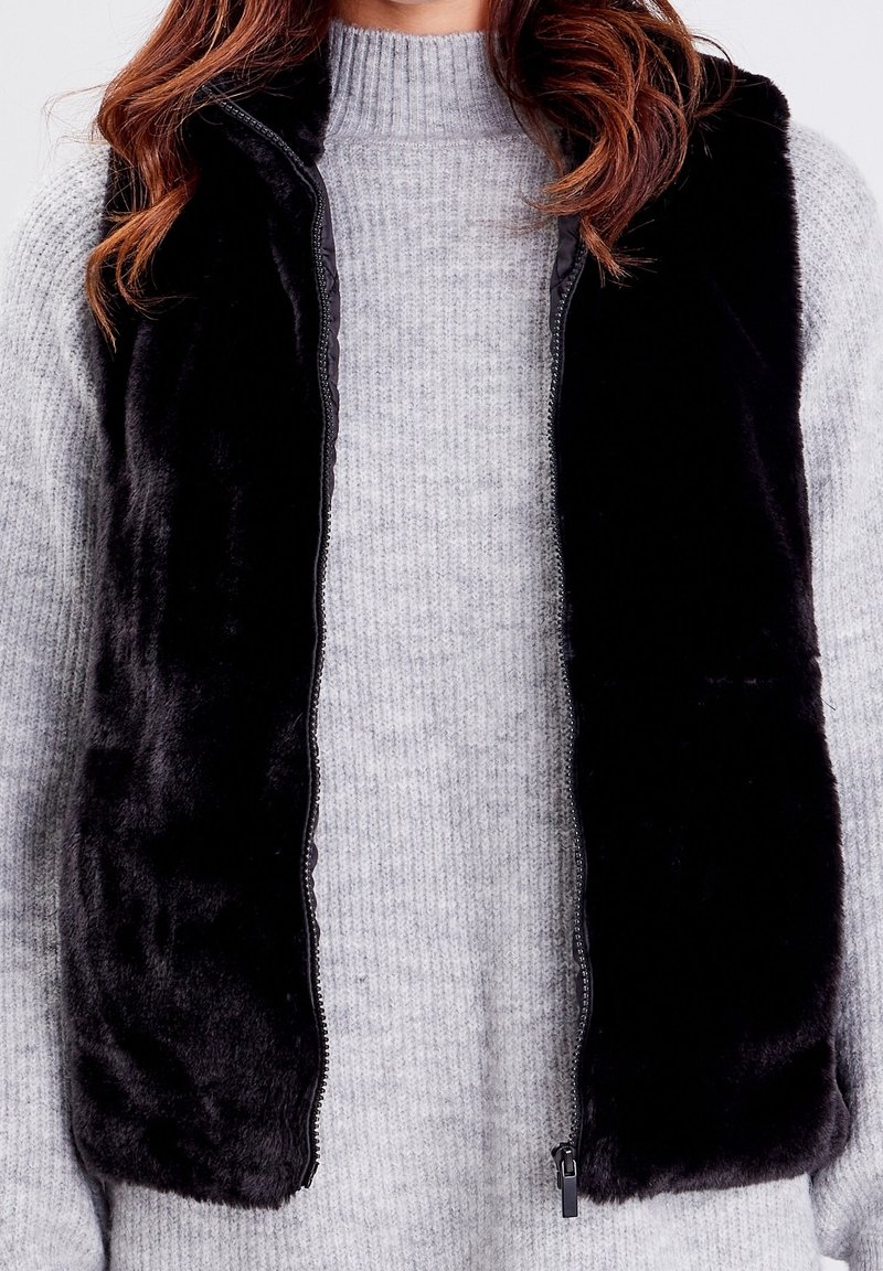 Personne portant une veste en fourrure noire par-dessus un pull en maille côtelée gris clair à manches longues et col montant.