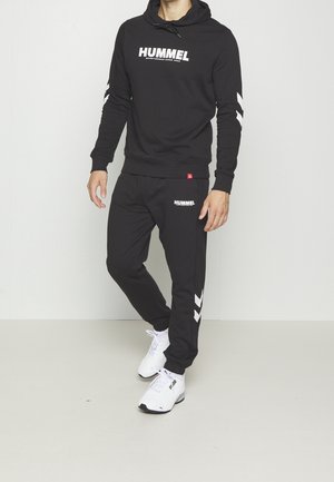 Mand iført sort Hummel hættetrøje og matchende joggers med hvide chevron-designs, kombineret med hvide Puma sneakers, stående indendørs.