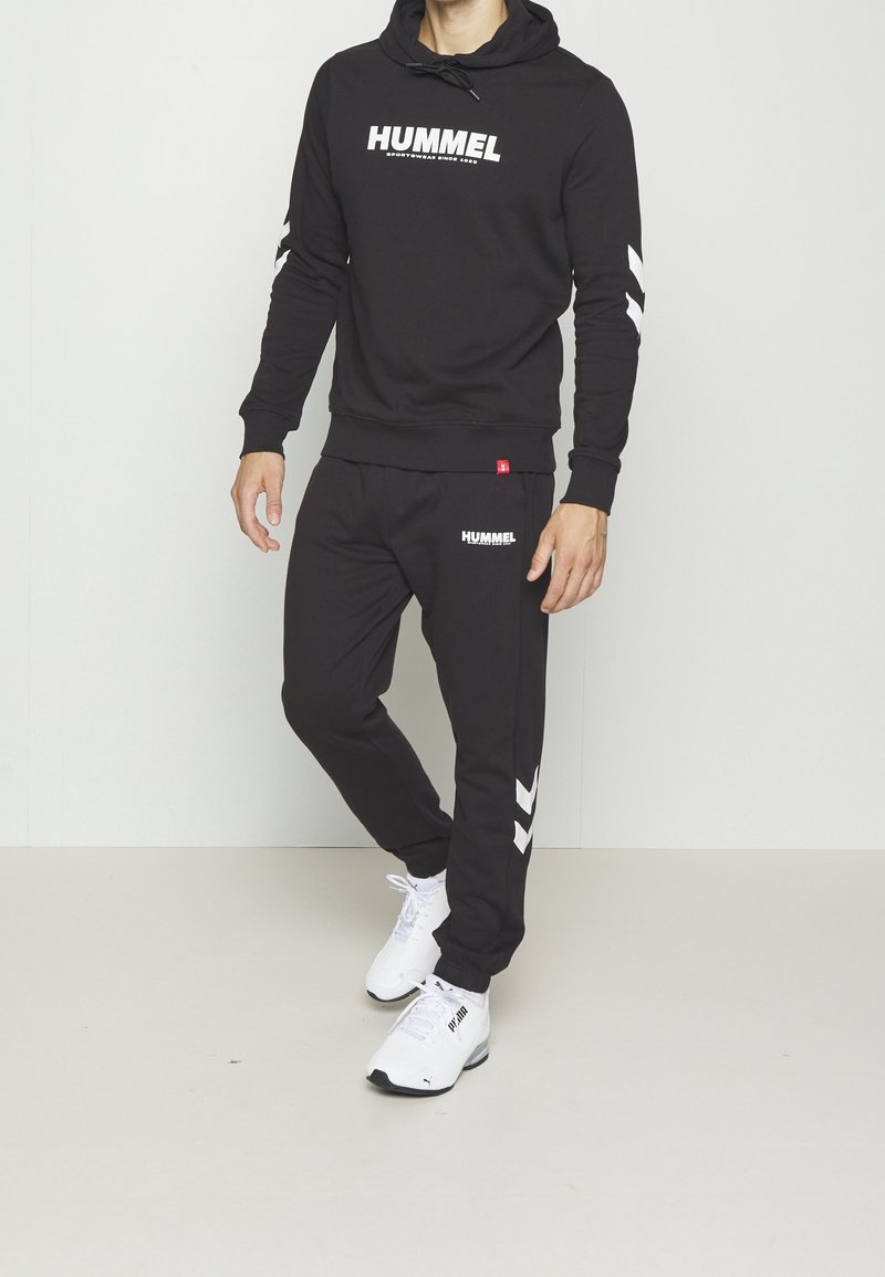 Homme portant un sweat à capuche noir Hummel et un jogging assorti avec des motifs chevrons blancs, associé à des baskets blanches Puma, debout à l'intérieur.