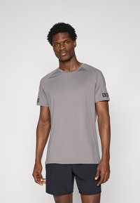 T-shirt de sport gris à manches courtes avec un col rond et un logo noir sur la manche gauche. Tissu lisse avec une texture légère.