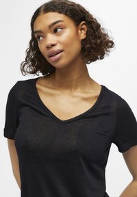 Zwarte v-hals t-shirt met korte opgerolde mouwen en een klein zakje. De stof lijkt lichtgewicht en licht doorschijnend met een zachte textuur.