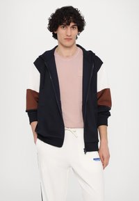 Navy zip-up hoodie with white and brown accents, layered over a light pink t-shirt. Comfortable fabric with elastic cuffs and waistband.
