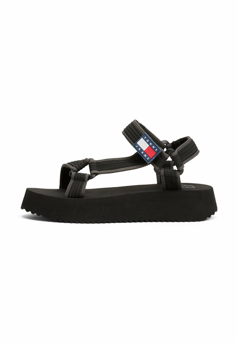 Sandalias de plataforma negras con tiras de tela entrelazadas, que presentan un parche de logo de marca en rojo, blanco y azul en el costado. Suela texturizada.