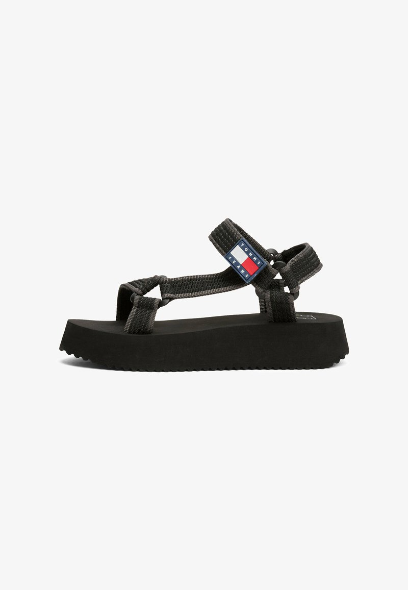 Sandalias de plataforma negras con tiras de tela entrelazadas, que presentan un parche de logo de marca en rojo, blanco y azul en el costado. Suela texturizada.