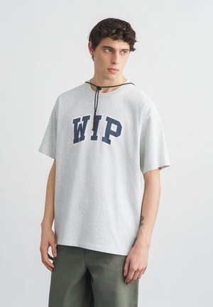 Młody mężczyzna w jasnoszarej koszulce z dużymi granatowymi literami „WIP” i ciemnozielonych spodniach, stojący na tle jasnego, jednolitego tła.