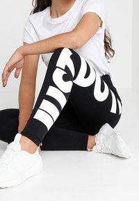 Osoba siedząca w pozycji skrzyżnej, ubrana w czarne legginsy z dużym białym napisem i białe tenisówki, z rękami opartymi na kolanach, w białej koszulce z krótkim rękawem.