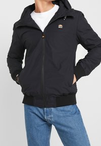 Svart zip-up jacka med huva, ribbade ärmslut och nederkant, med en liten logotyp på bröstet. Slät tyg med sidofickor.