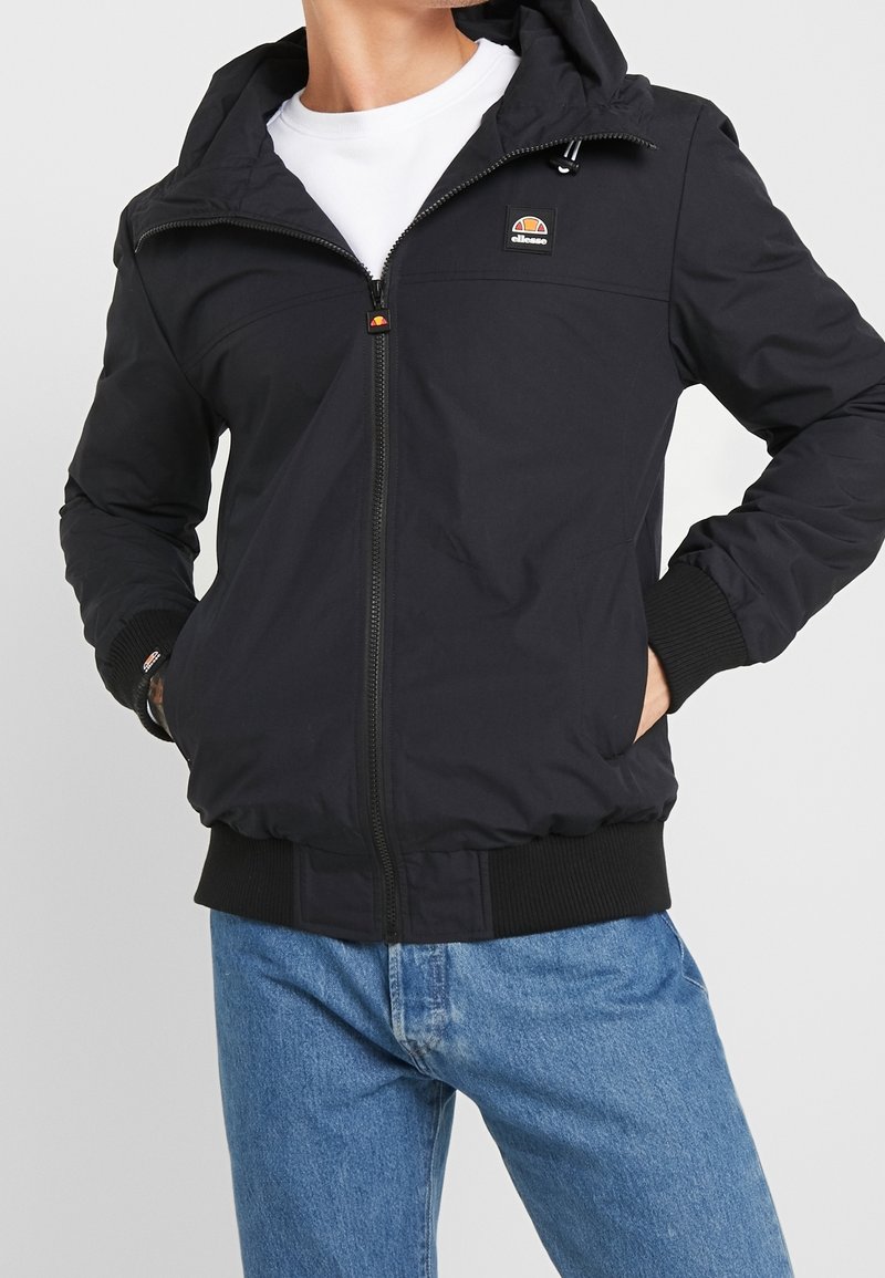 Svart zip-up jacka med huva, ribbade ärmslut och nederkant, med en liten logotyp på bröstet. Slät tyg med sidofickor.