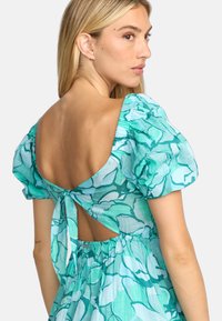 Robe à manches courtes, de couleur aqua avec des fleurs, présentant une découpe profonde au dos et un détail à nouer. Fabriquée dans un tissu léger avec une apparence texturée.
