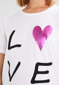 Camiseta blanca de algodón con texto negro que dice "AMOR" y un diseño de corazón fucsia brillante. Mangas cortas y cuello redondo.