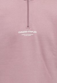 Gaiši rozā ceturkšņa rāvējslēdzēja džemperis no mīksta auduma. Uz krūtīm ir balts uzraksts "Curated Staples" un citi zīmola elementi.