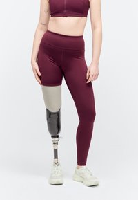 Personne portant une tenue de sport bordeaux avec une jambe gauche prothétique et des baskets blanches, debout devant un fond clair uni.