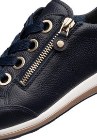 Tmavě modrá kožená teniska s tmavě modrými tkaničkami, zlatým zipem a otvory na tkaničky, bílou a hnědou podrážkou, detailní záběr z boku.