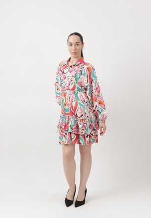 Vrouw staat tegen een witte achtergrond, gekleed in een kleurrijke, lange mouwen jurk met patroon en ruches, en zwarte hoge hakken.