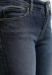 Zbliżenie na ciemnoniebieskie dżinsy w okolicy pasa i górnej części uda, z widoczną przednią kieszenią, szlufką na pasek oraz detalami szycia.