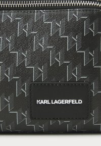 Svart lommebok med et teksturert mønster av sammenflettede K-er, sølvfarget glidelås og en svart lærlabel med hvit merkevare. 