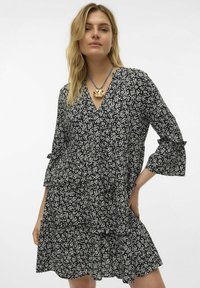 Vestido negro con estampado floral blanco, escote en V, diseño en capas y mangas de campana. Lleva un collar de péndulo dorado y un cordón azul.