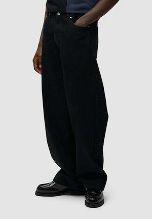 Persona che indossa pantaloni neri larghi con la mano in tasca e scarpe nere lucide, in piedi davanti a uno sfondo neutro.