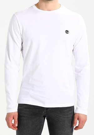 Homme portant un t-shirt blanc uni à manches longues et col rond avec un petit logo d'arbre noir sur la poitrine gauche, associé à un jean foncé.