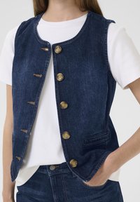 Sötétkék farmermellény kerek nyakkivágással, öt sárgaréz gombbal záródik, kontrasztos varrásokkal és egy kis oldalzsebből áll.