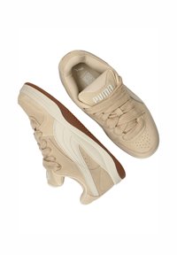 Beige Puma sneakers met dikke veters, witte zijstrips, bruine zoolaccenten, en het Puma-logo op de tong en inlegzool.