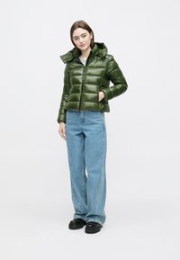 Grøn dunjakke med skinnende finish, lynlåslukning foran og hætte. Modellen bærer lyseblå jeans med vide ben og sort fodtøj.