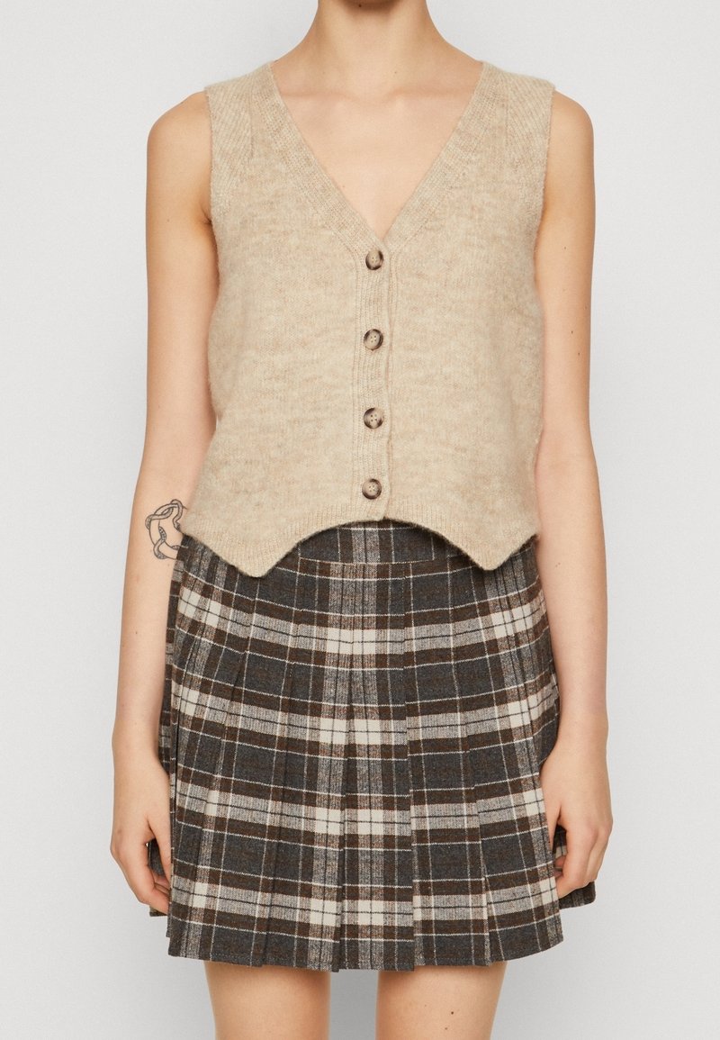 Gilet beige lavorato a maglia con scollo a V, caratterizzato da cinque bottoni marroni e un orlo a ondine, abbinato a una gonna plissettata a quadri marroni e crema.