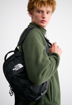 Sac à dos noir avec logo, fabriqué en nylon ; doté de bretelles rembourrées et de cordes élastiques à l'avant. Porté sur une veste en polaire verte olive.