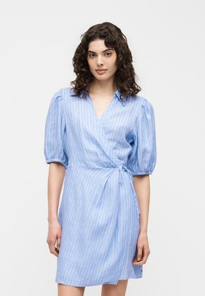 Γυναίκα με σγουρά σκοτεινά μαλλιά φοράει ανοιχτό μπλε φόρεμα με κοντά μανίκια και λεπτές κάθετες ρίγες, στέκεται μπροστά σε λευκό φόντο.