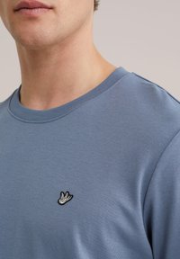Close-up van een persoon die een blauw shirt met ronde hals draagt, met een klein geborduurd wit-zwart vogellogo op de borst.