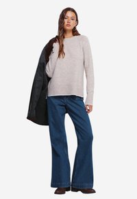 Maglione knit color beige chiaro con scollo a girocollo, abbinato a jeans a gamba larga blu scuro. Il modello tiene un giubbotto scuro e indossa stivali marroni.