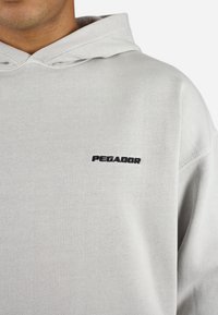 Lichtgrijze hoodie met ribbeltextuur, voorzien van een zwart geborduurd logo "PEGADOR" op de linkerkant van de borst. Capuchont ontwerp met een relaxte pasvorm.