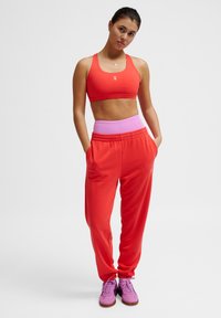 Rode sportbeha, rode joggingsbroek met roze tailleband, stof is glad met een losse pasvorm, heeft zakken, gecombineerd met roze sneakers.