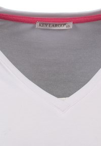 Camiseta blanca de cuello en V con ribete rosa alrededor del escote, hecha de una tela suave. La etiqueta dice "KEY LARGO" y talla XS.