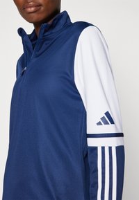 Marineblå kvart-zip trøje lavet af glat materiale. Har hvide ærmer med tre striber og logo på skulderen.